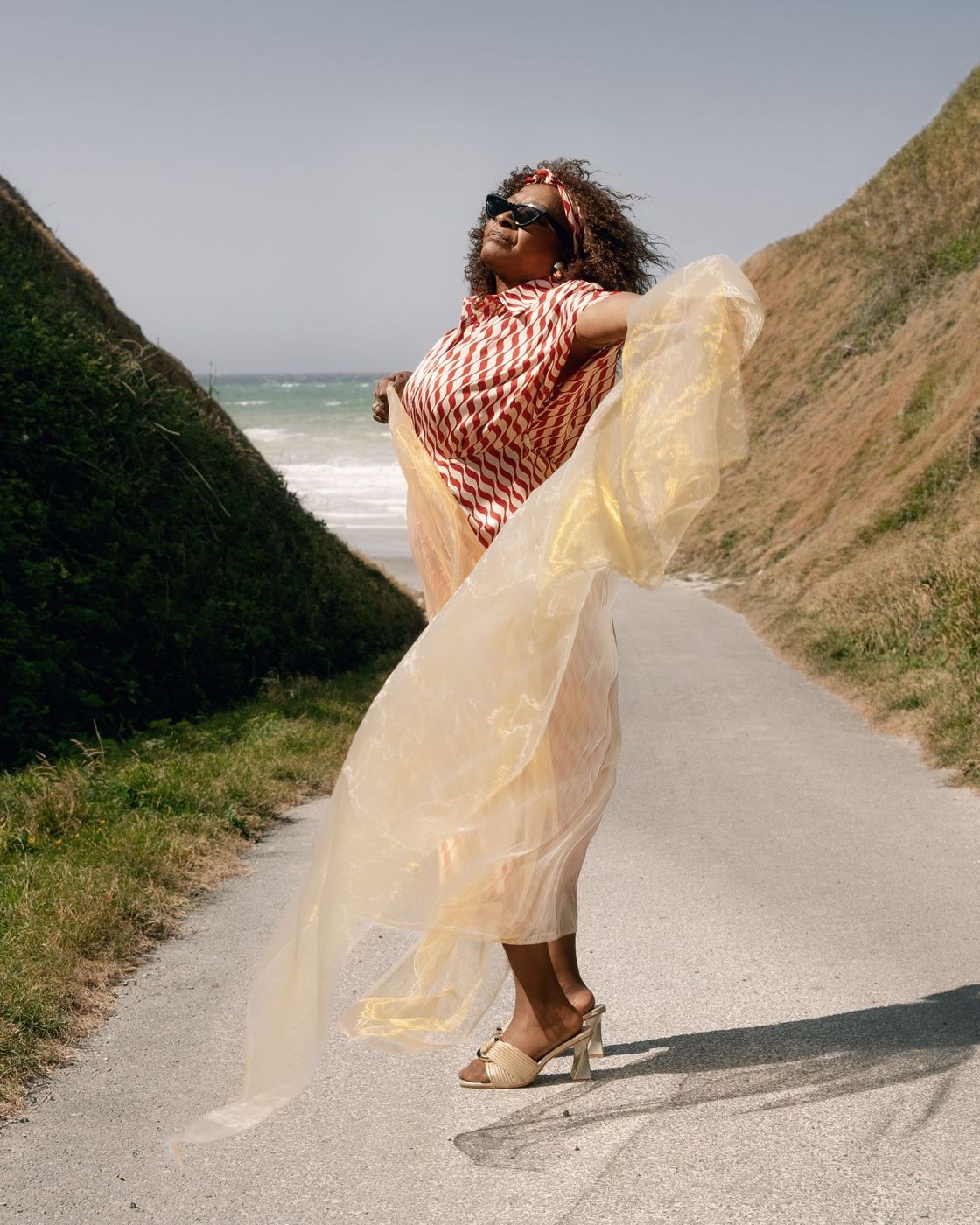 Linda Lee Hopkins en robe rayée, un voile doré au vent, sur un chemin face à la mer